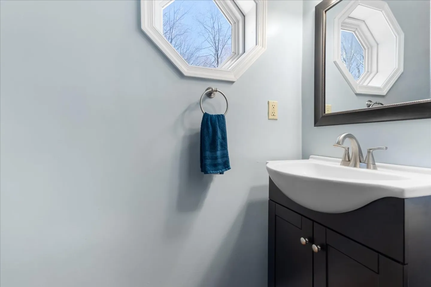 Bathroom vanity with dark cabinet, white sink, brushed metal faucet, framed mirror, and an octagonal window on light blue walls.