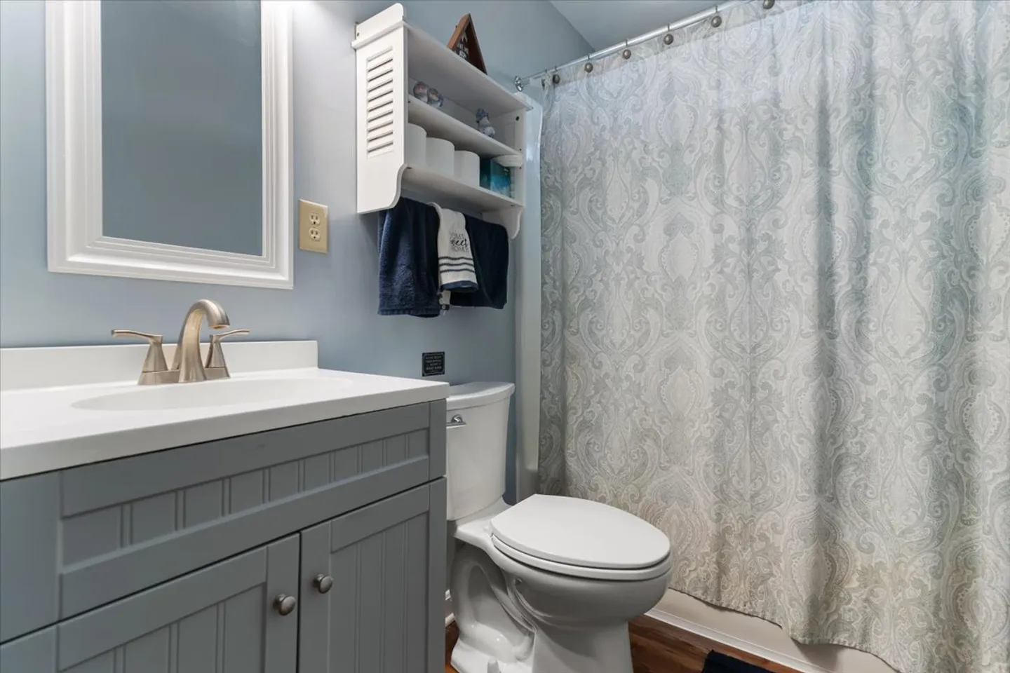 Bathroom with a grey vanity, white framed mirror, toilet, wall shelf with towels, and a patterned shower curtain.