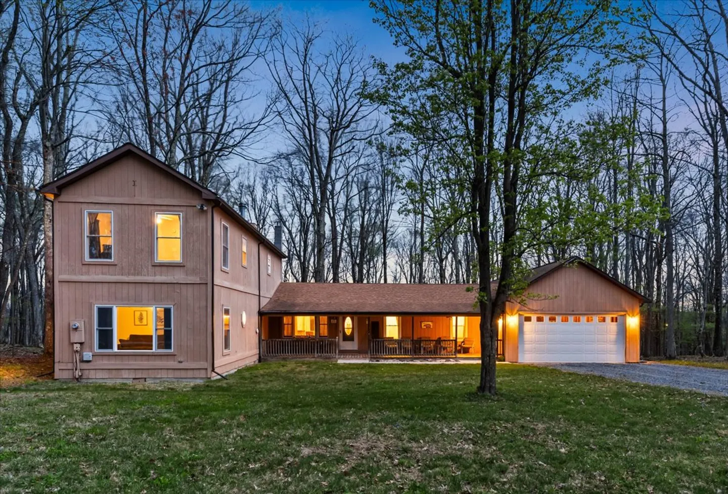 Exterior dusk view of a two-story tan siding home with attached garage, covered front porch, and lawn surrounded by bare trees.