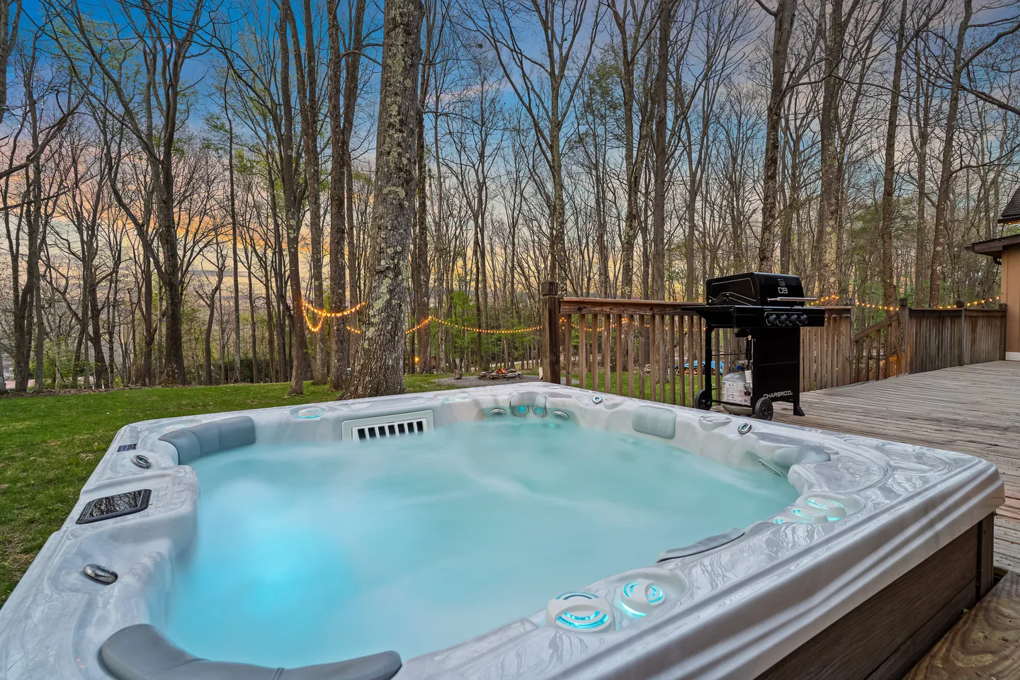 An outdoor hot tub with illuminated water next to a wooden deck with a barbecue grill and wooded view.