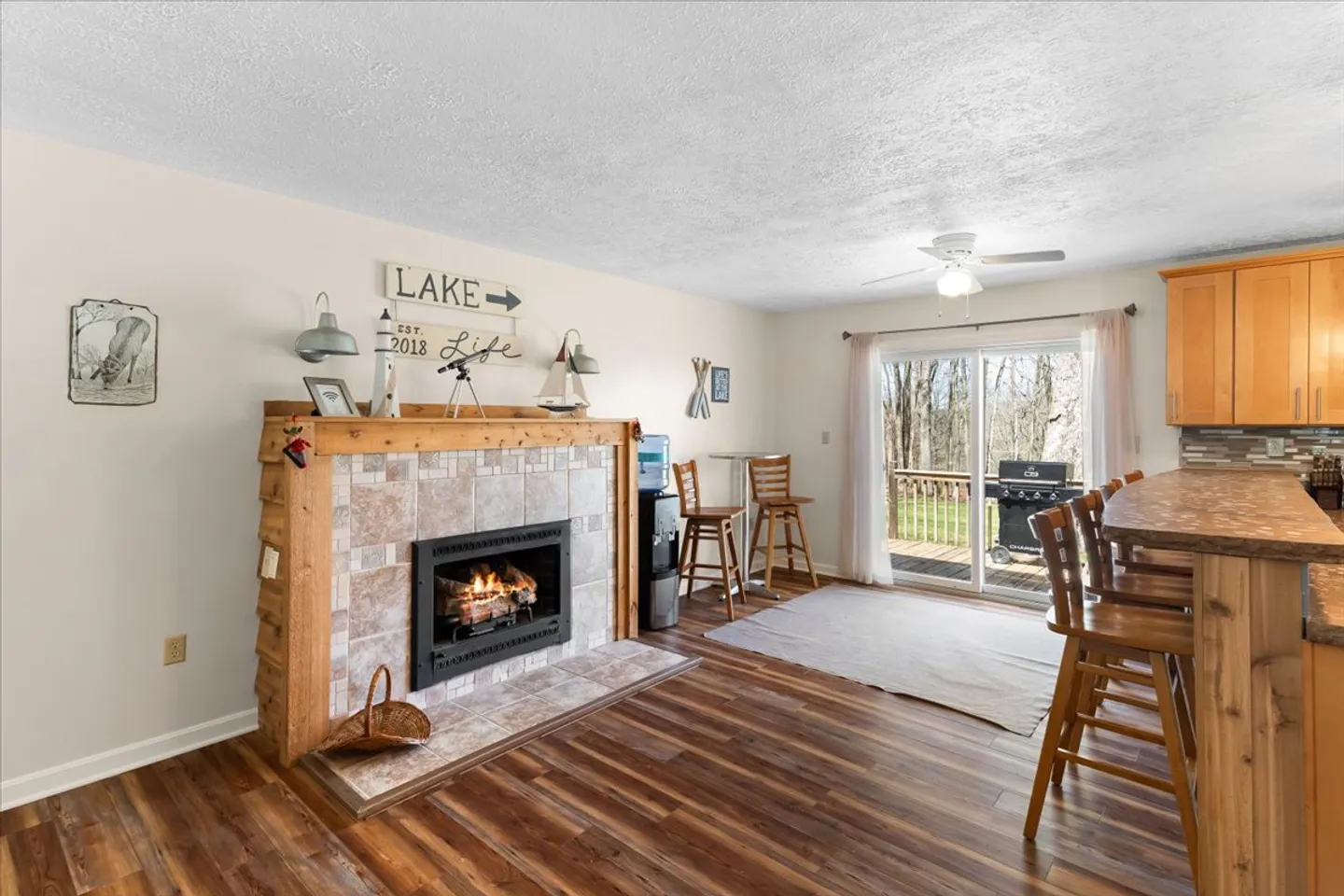 Open living and dining area featuring a tile-surround fireplace with a wooden mantel, wood-look flooring, breakfast bar seating, and sliding glass doors leading to an outdoor deck.