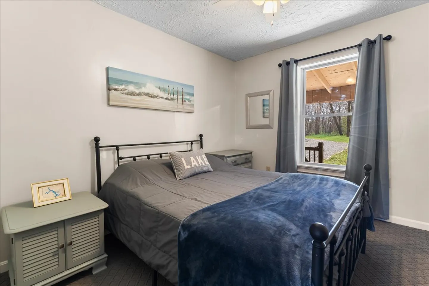 Bedroom with a black metal frame bed, gray bedding, two painted nightstands, and a window looking out toward a covered porch and trees.