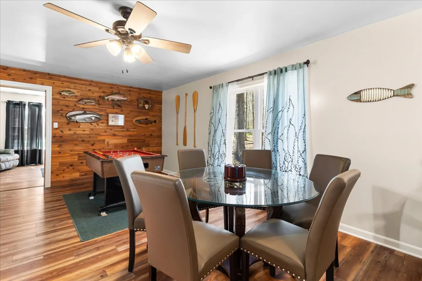 Dining area with a round glass table, upholstered chairs, and a rod hockey table set against a wood-paneled accent wall with fish decor.