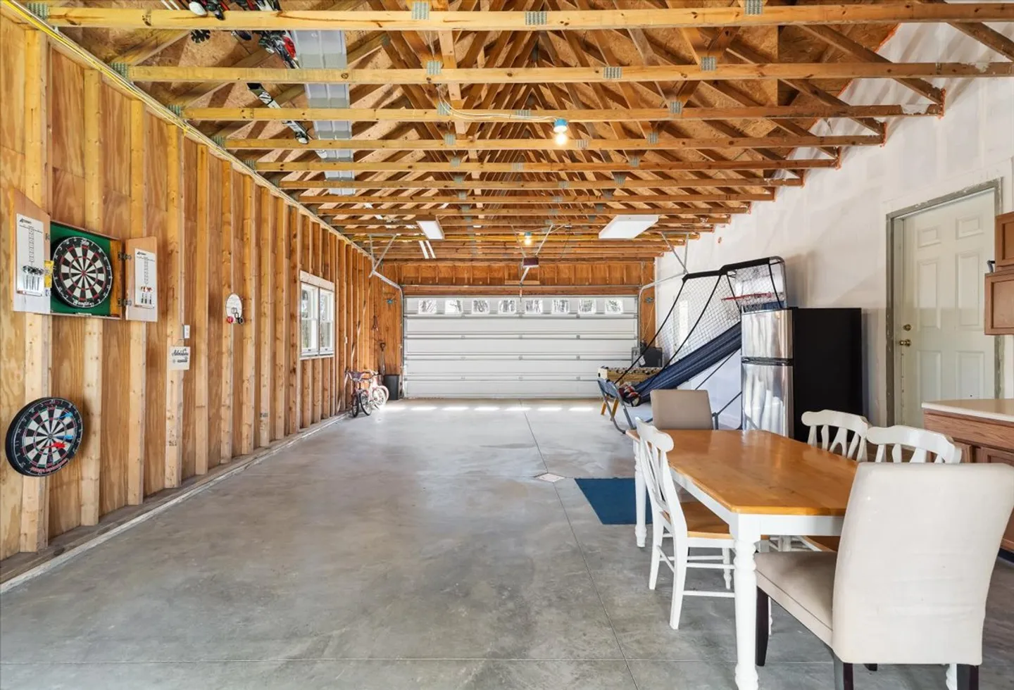 Spacious garage with exposed wooden rafters, concrete floor, dartboards, basketball arcade game, and a dining table with chairs.