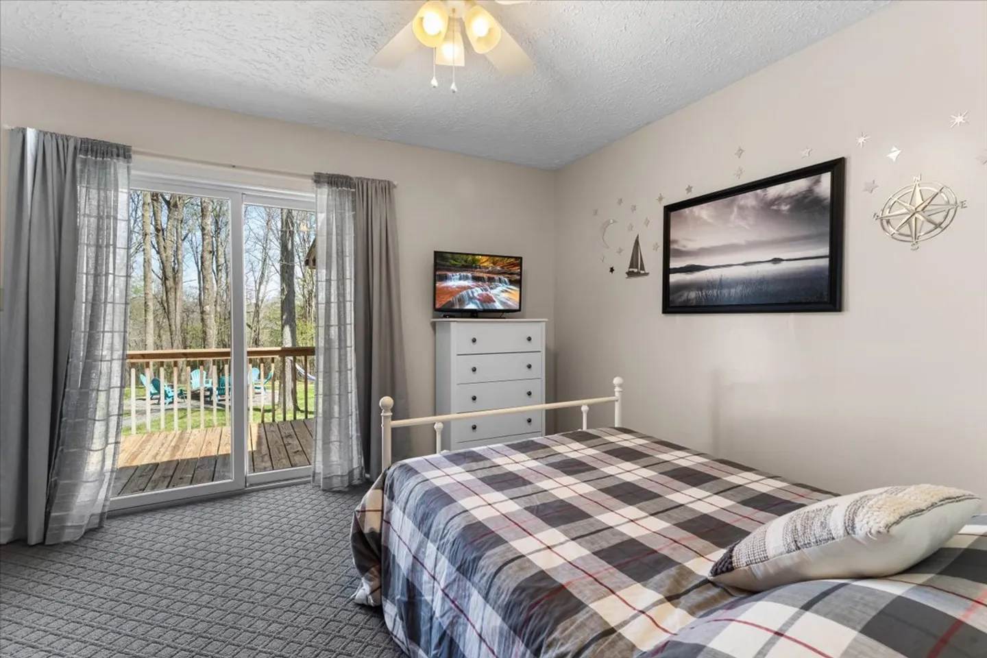 Bedroom with a plaid bedspread, white dresser with TV, textured carpet, and sliding glass doors leading to an outdoor deck.