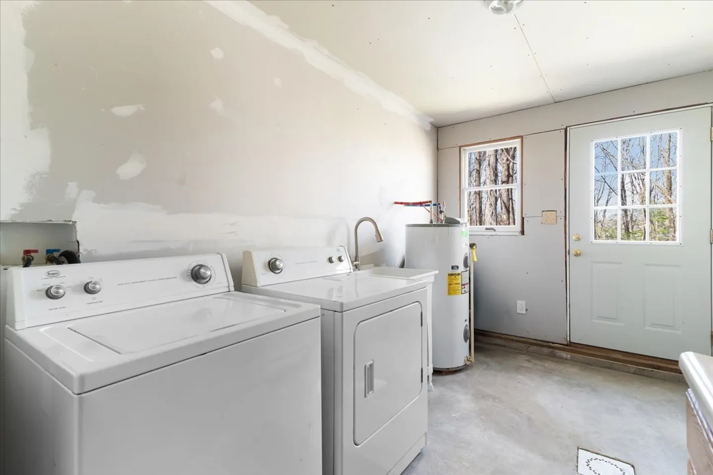 A utility room with a white washing machine, dryer, utility sink, water heater, concrete floor, and an exterior door.