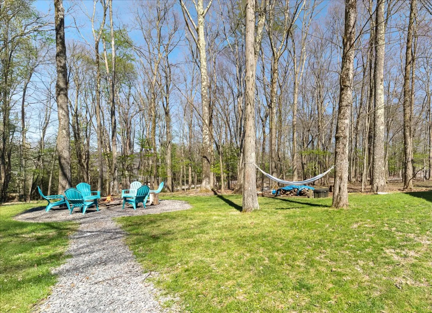 Outdoor yard with a gravel path leading to a fire pit with turquoise Adirondack chairs, alongside a hammock tied between trees in a wooded area.