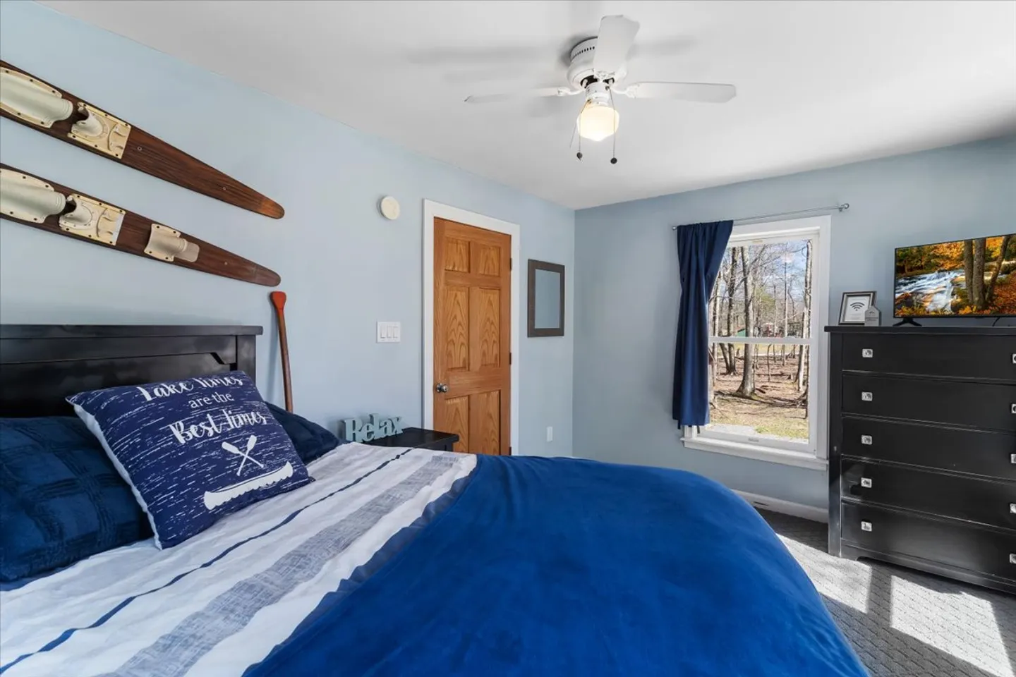 Bedroom with blue walls, a bed made with blue bedding, rustic water ski wall decor, a window, a ceiling fan, and a black dresser with a television.