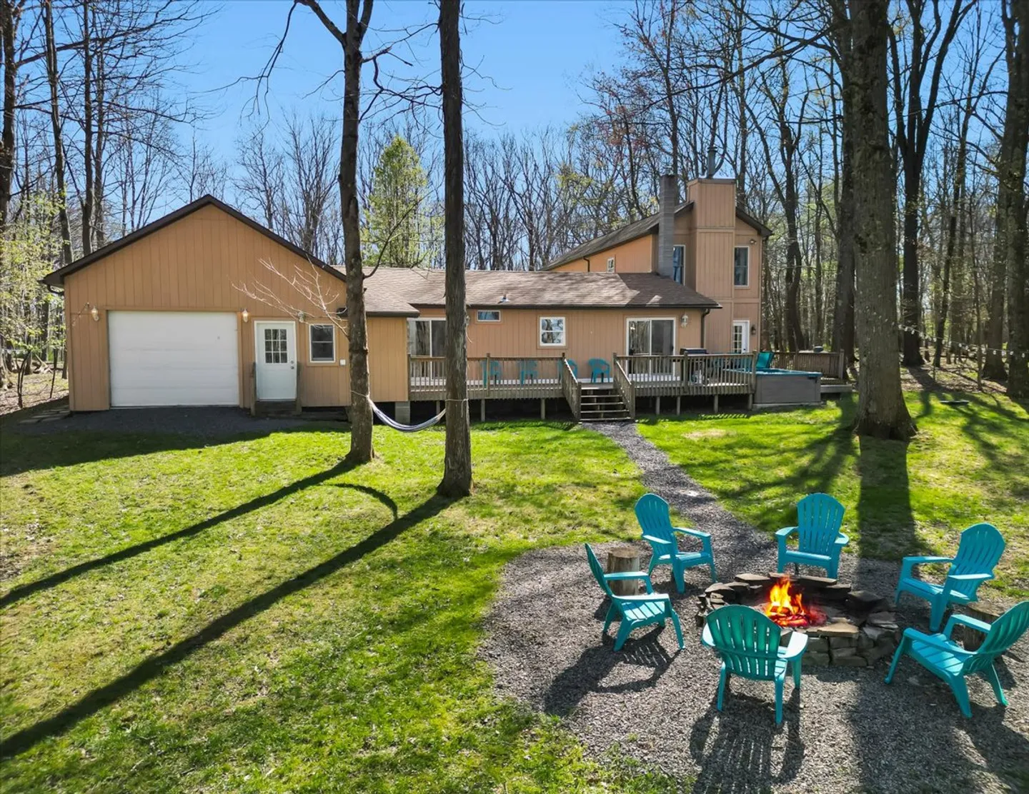 Rear exterior of a light brown house featuring an elevated wooden deck, garage, fire pit with bright turquoise Adirondack chairs, and a hammock in a wooded yard.