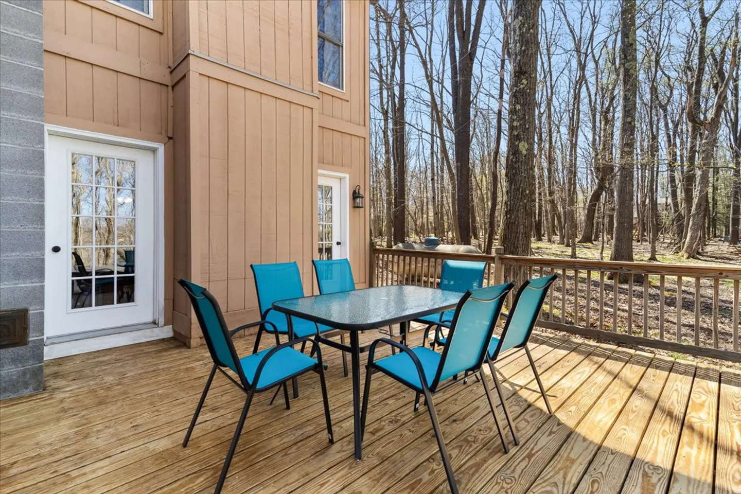 Wooden deck with a glass-topped dining table and bright blue chairs surrounded by tall bare trees.