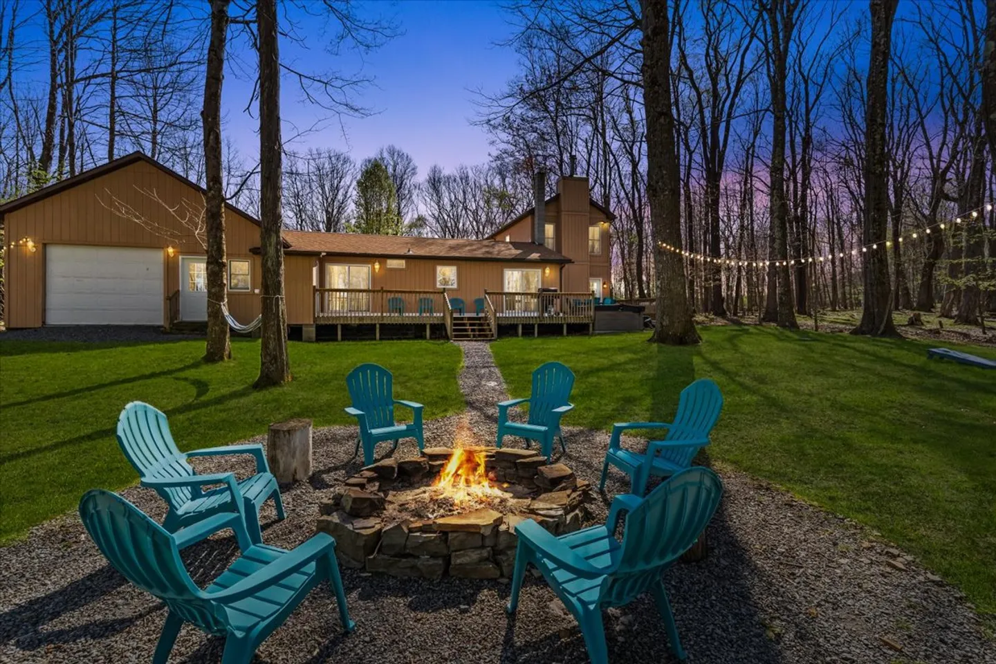 A backyard fire pit with burning wood surrounded by turquoise Adirondack chairs, with a stone path leading toward a wood-sided house and deck in a wooded yard at twilight.
