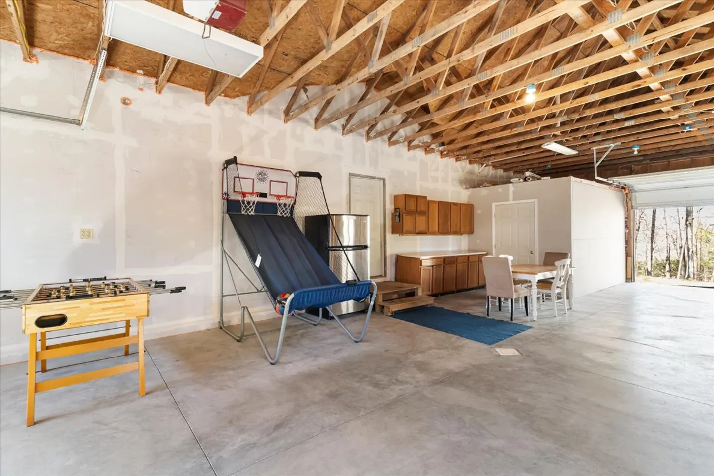 A spacious garage converted into a game room with concrete flooring, exposed truss rafters, an arcade basketball game, a foosball table, a refrigerator, and a dining table set.