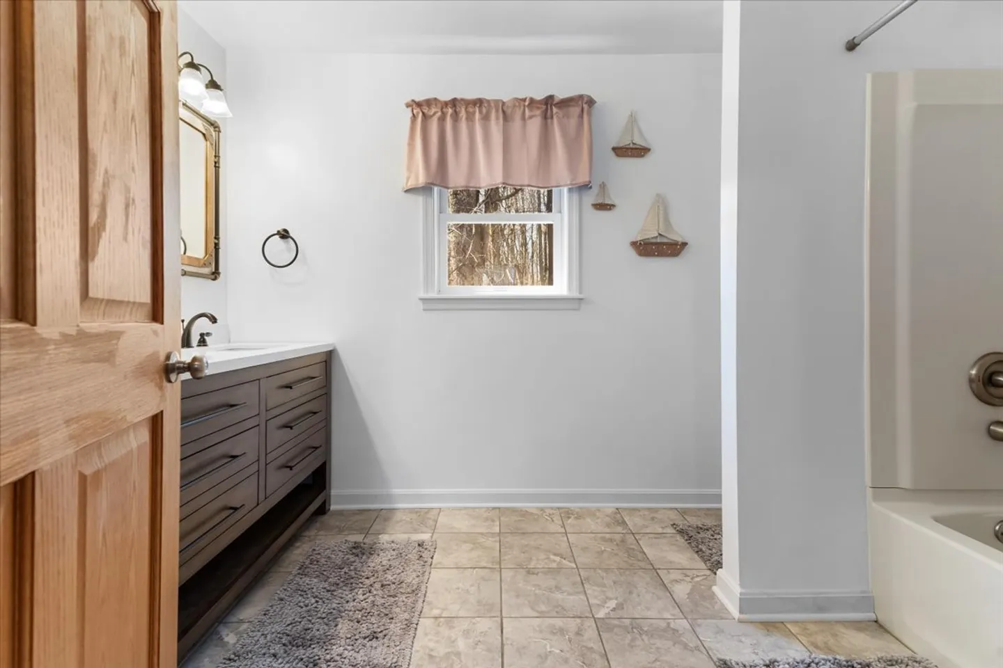 Bathroom with tile floor, dark vanity, mirror, window with curtain, sailboat wall decor, and bathtub shower surround.