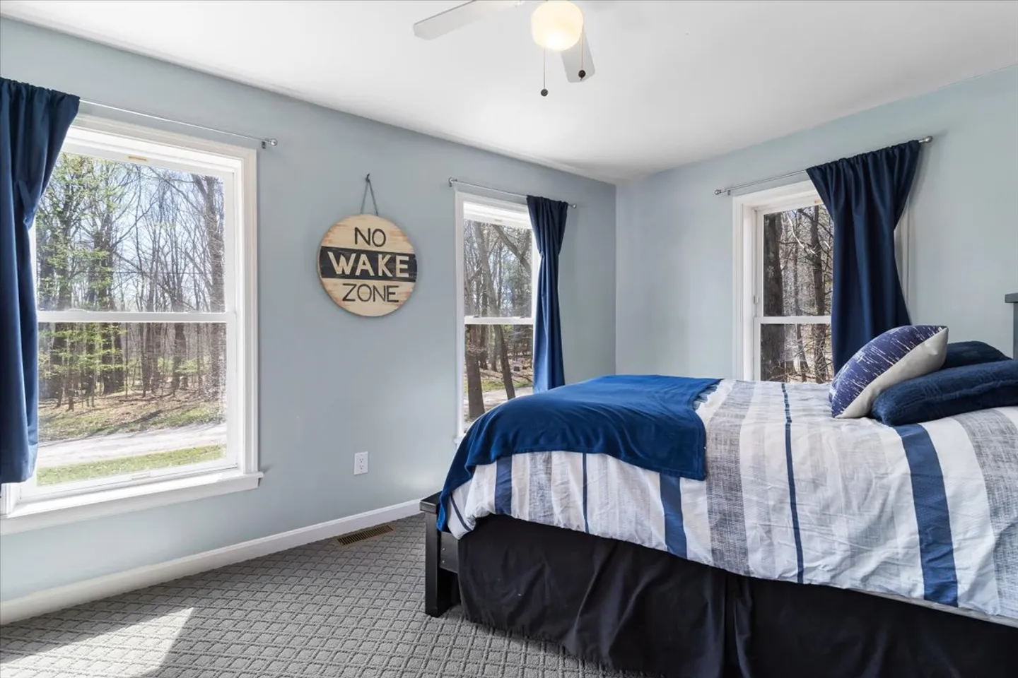 Bedroom with blue walls, carpeted floor, ceiling fan, multiple windows, and a bed with blue and white bedding.