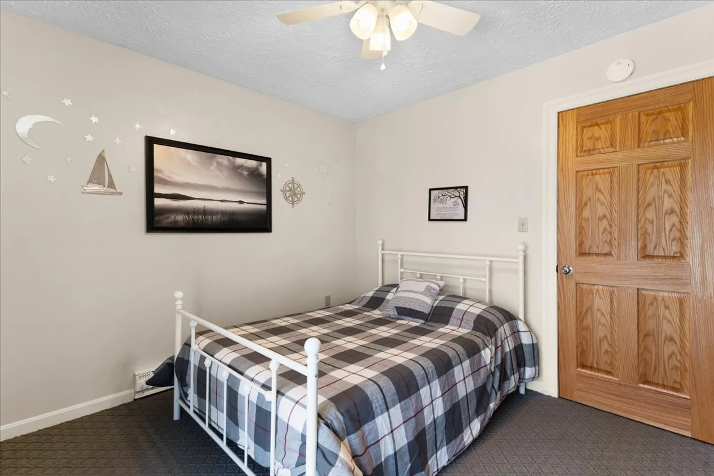 Bedroom featuring a white metal bed with a plaid comforter, carpeted floor, ceiling fan, and framed artwork on light-colored walls.
