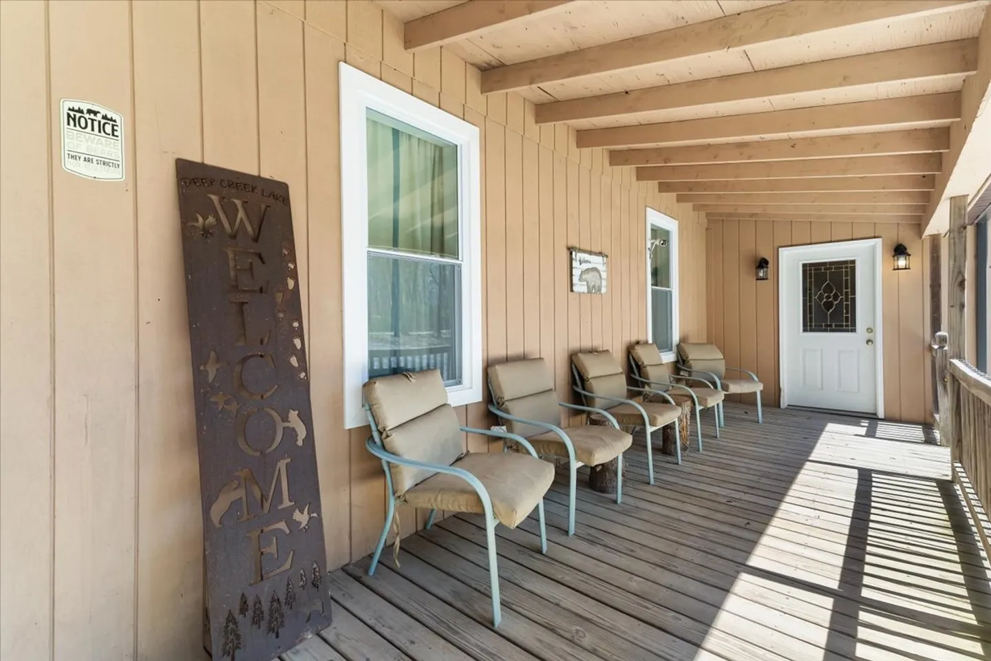 Covered porch with wood plank flooring, tan siding, and a row of padded chairs leading to an entry door.