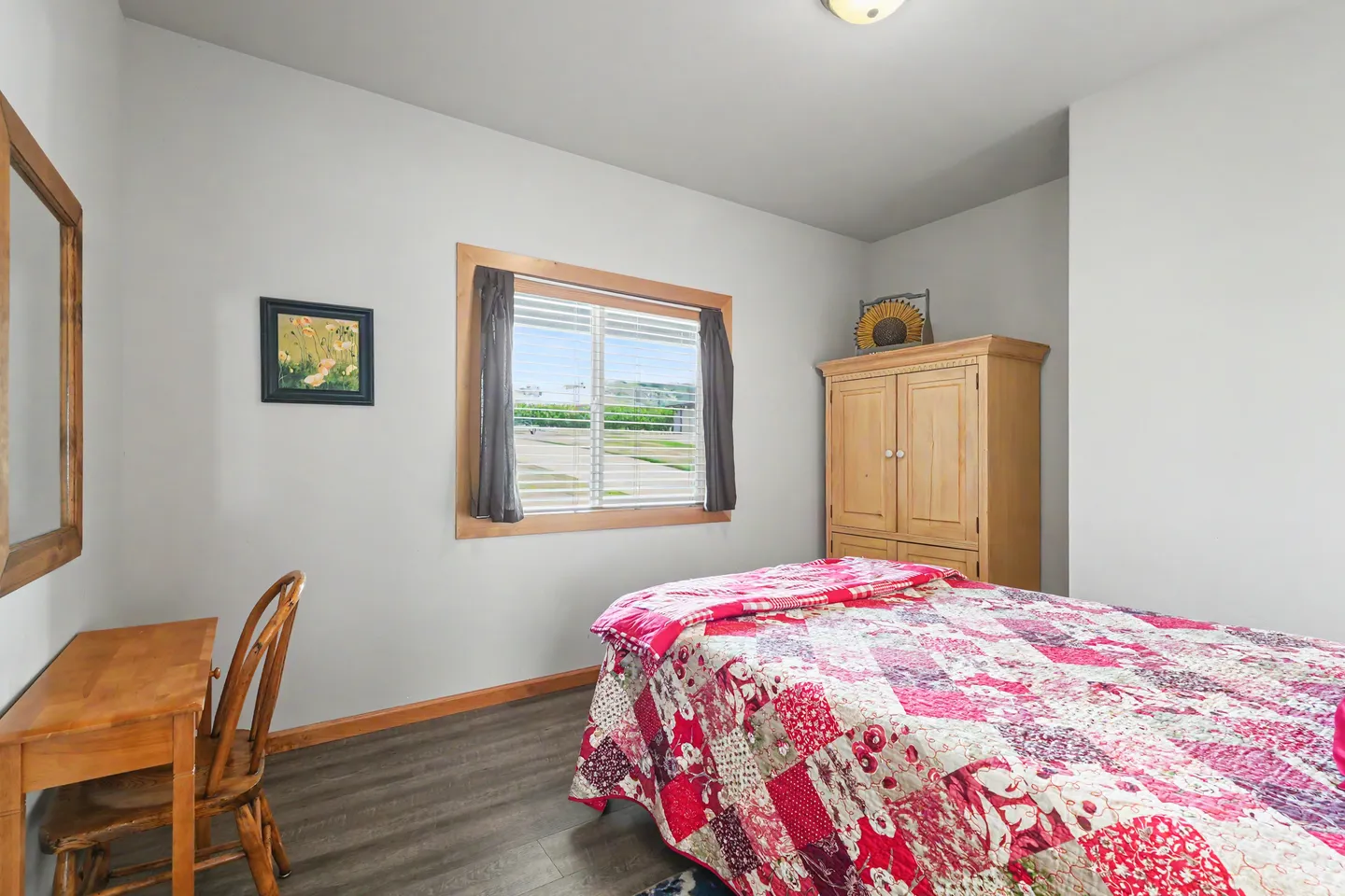 Bedroom featuring a bed with a red patchwork quilt, wood desk and chair, wooden armoire, and a window with blinds.