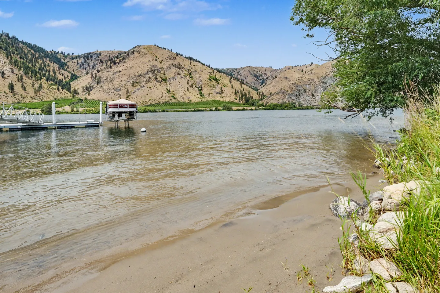 A sandy shoreline and river with a boat dock, boat lift, and arid rolling hills across the water under a clear blue sky.