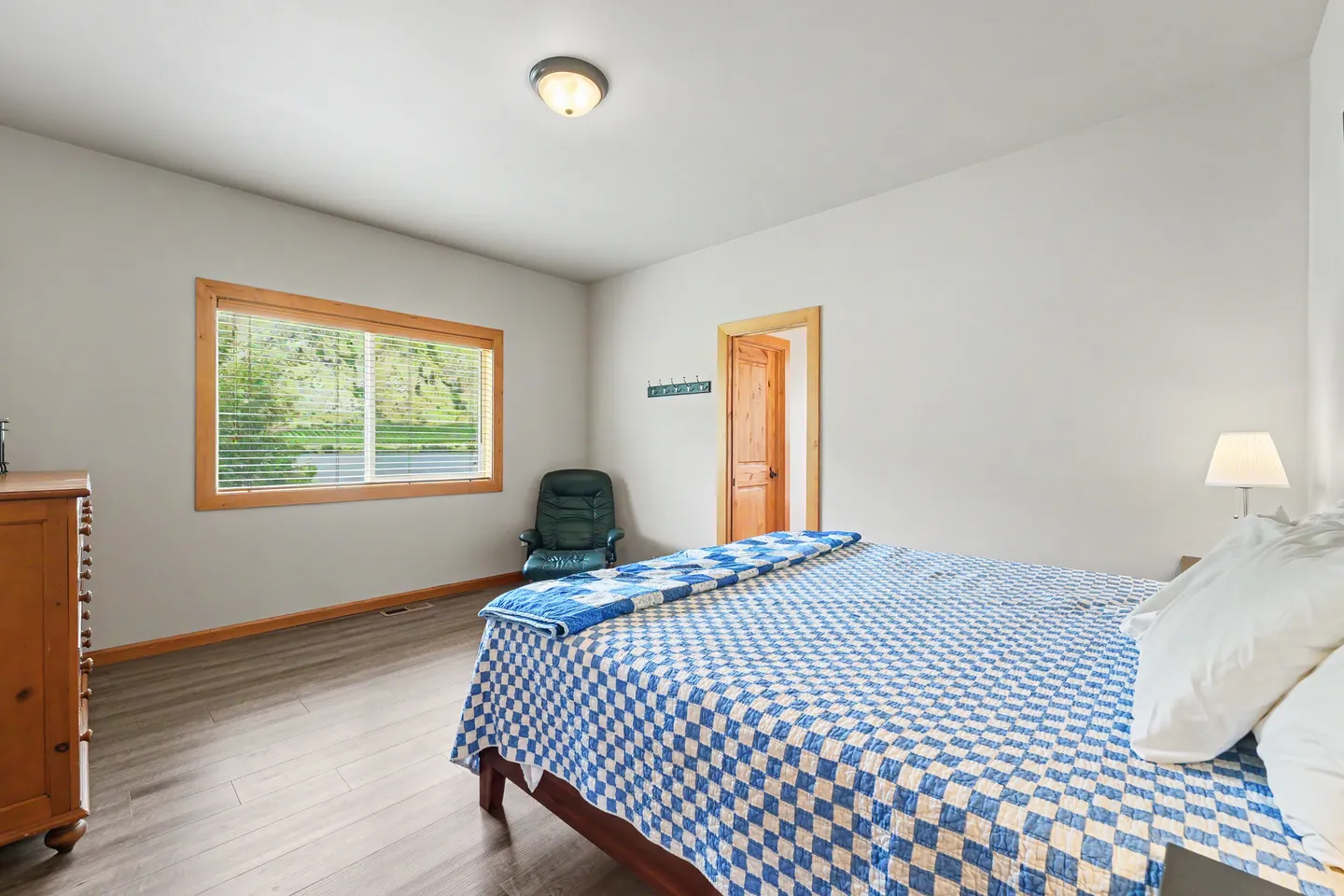 Bedroom with a bed made with a blue and white patterned quilt, a window looking outside, a dresser, and an armchair in the corner.