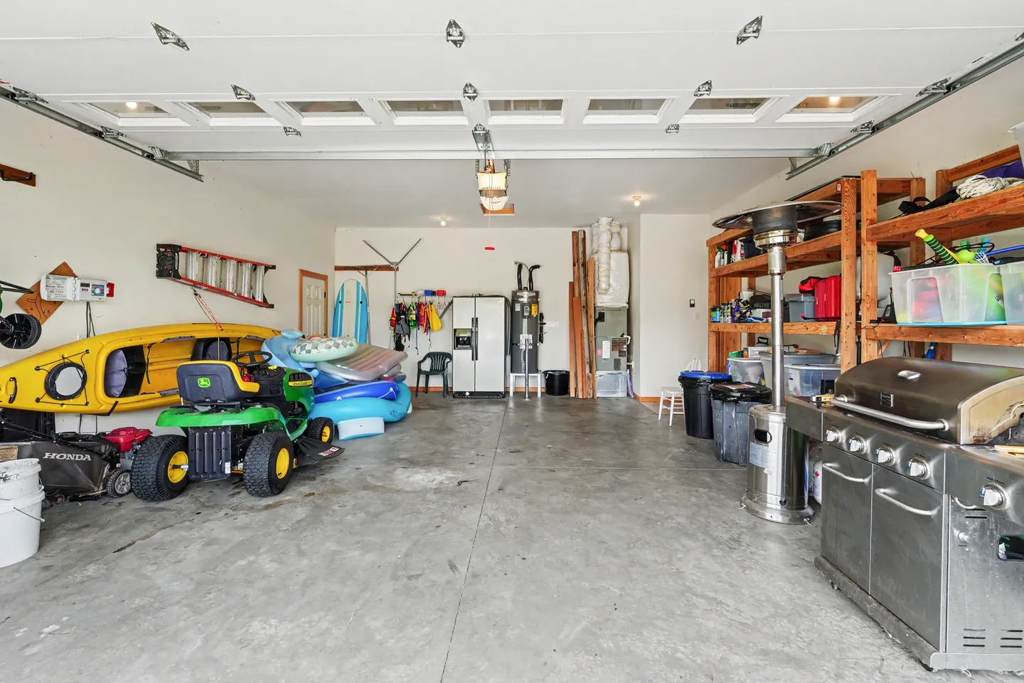 Spacious garage with concrete floor containing kayaks, riding lawn mower, barbecue grill, patio heater, and storage shelves.