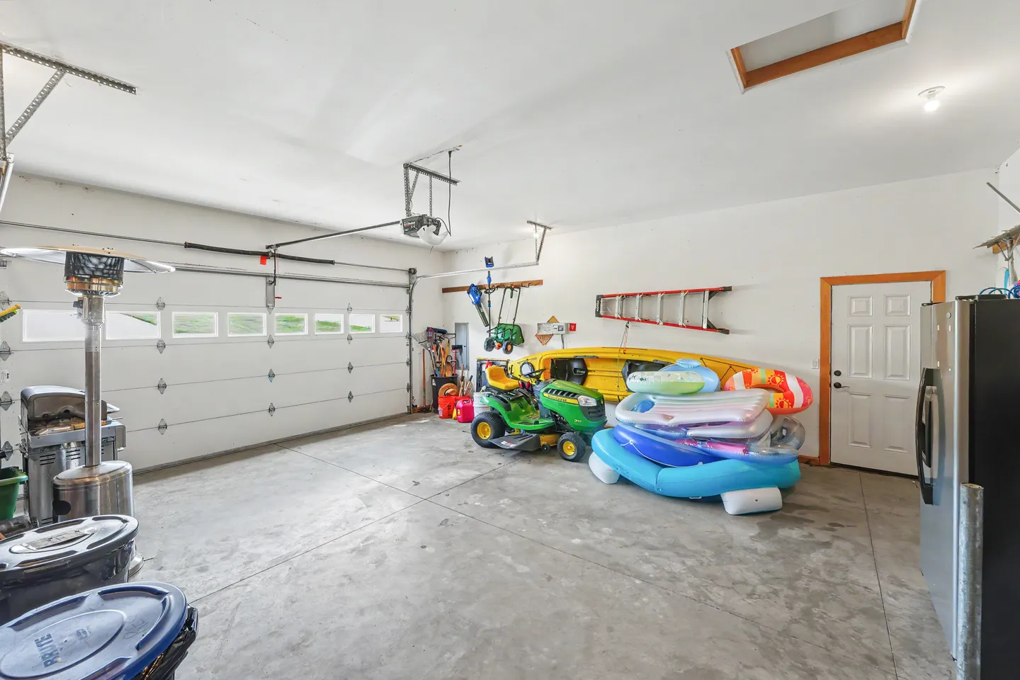 Interior view of an attached garage with concrete flooring, a closed garage door, lawn equipment, and inflatable pool floats.