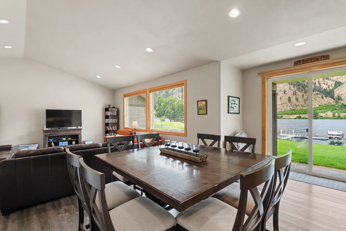 Dining area with a dark wooden square table and chairs adjacent to a living room with sliding glass doors opening to a river and mountain view.