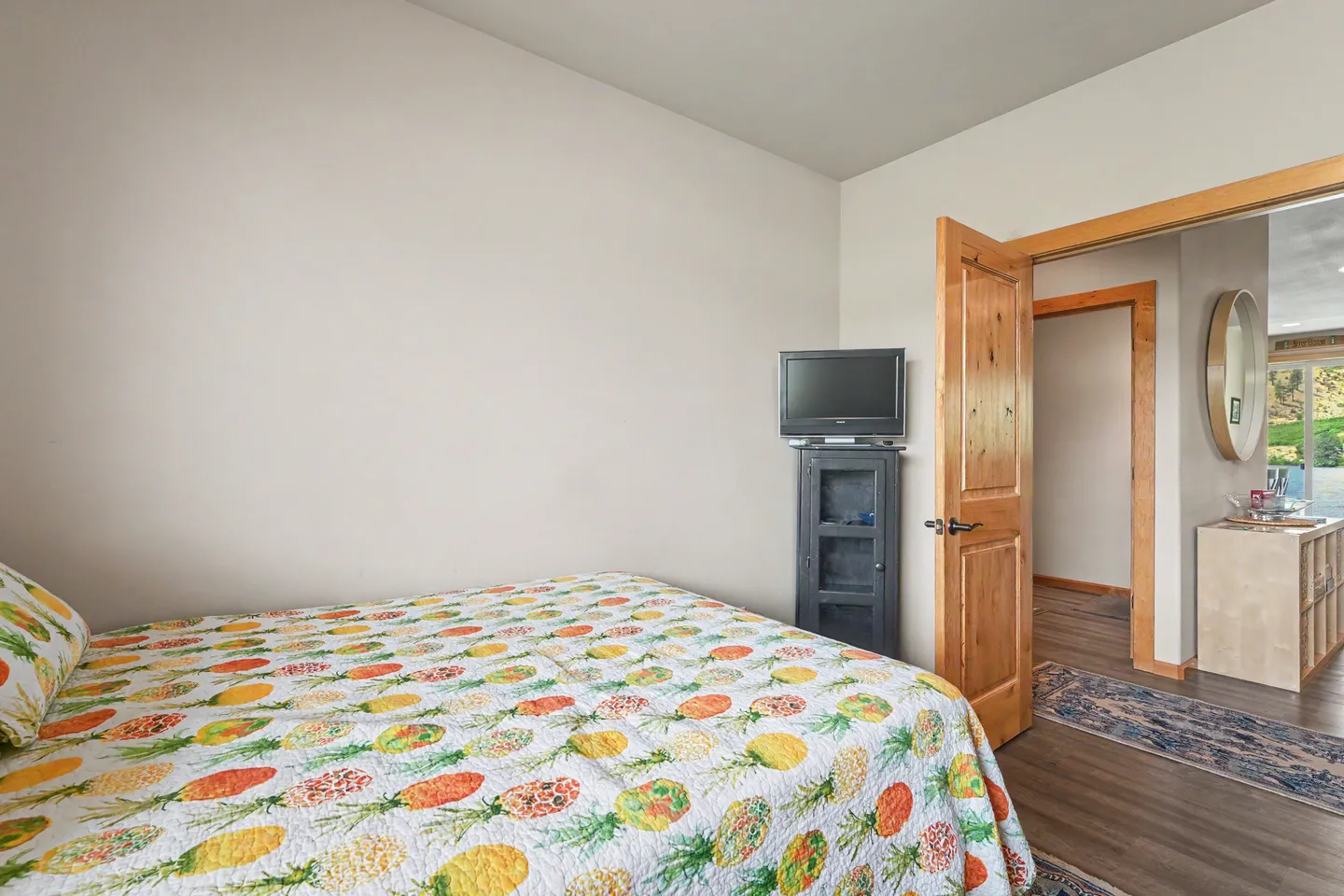 Bedroom with a bed made with a pineapple-patterned quilt, a television on a small stand, and an open wood door looking into a hallway.