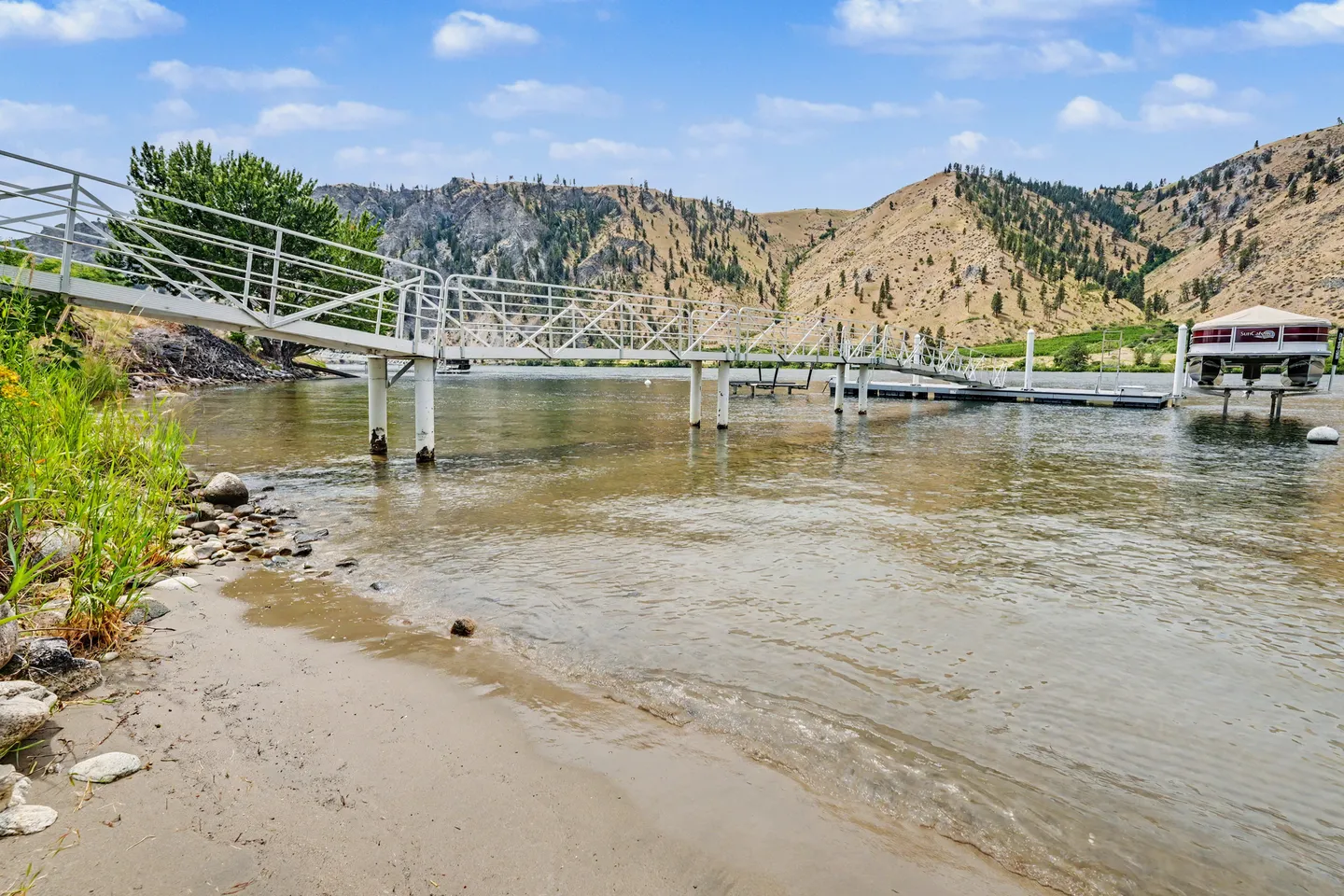A long metal pier and floating dock extending into a lake from a sandy shore with arid, mountainous hills across the water.