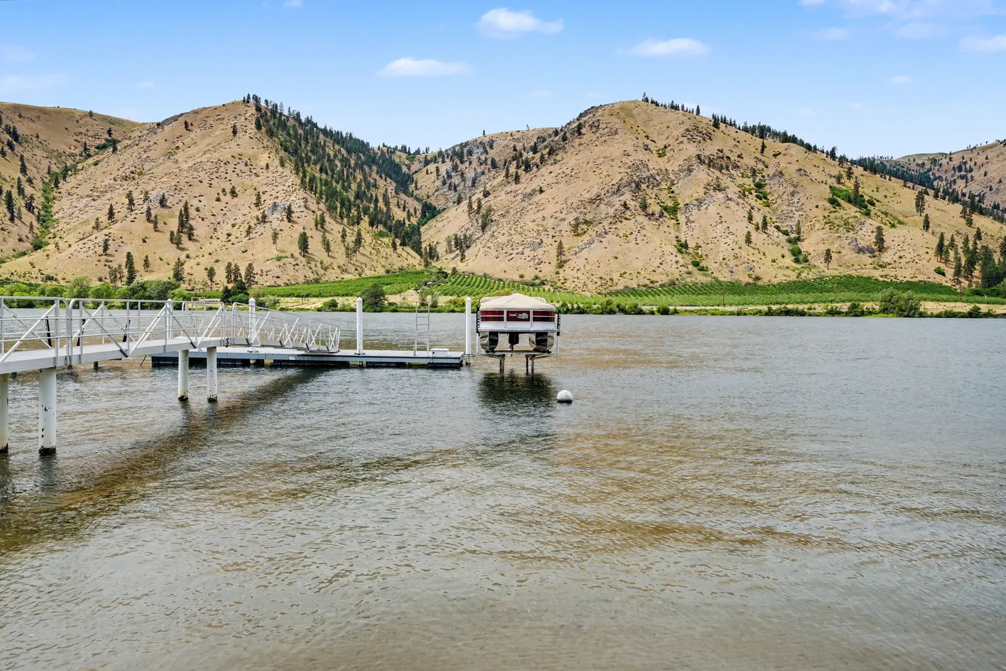 A floating boat dock with a covered pontoon boat on a lift extending into a lake against dry, rolling mountains.