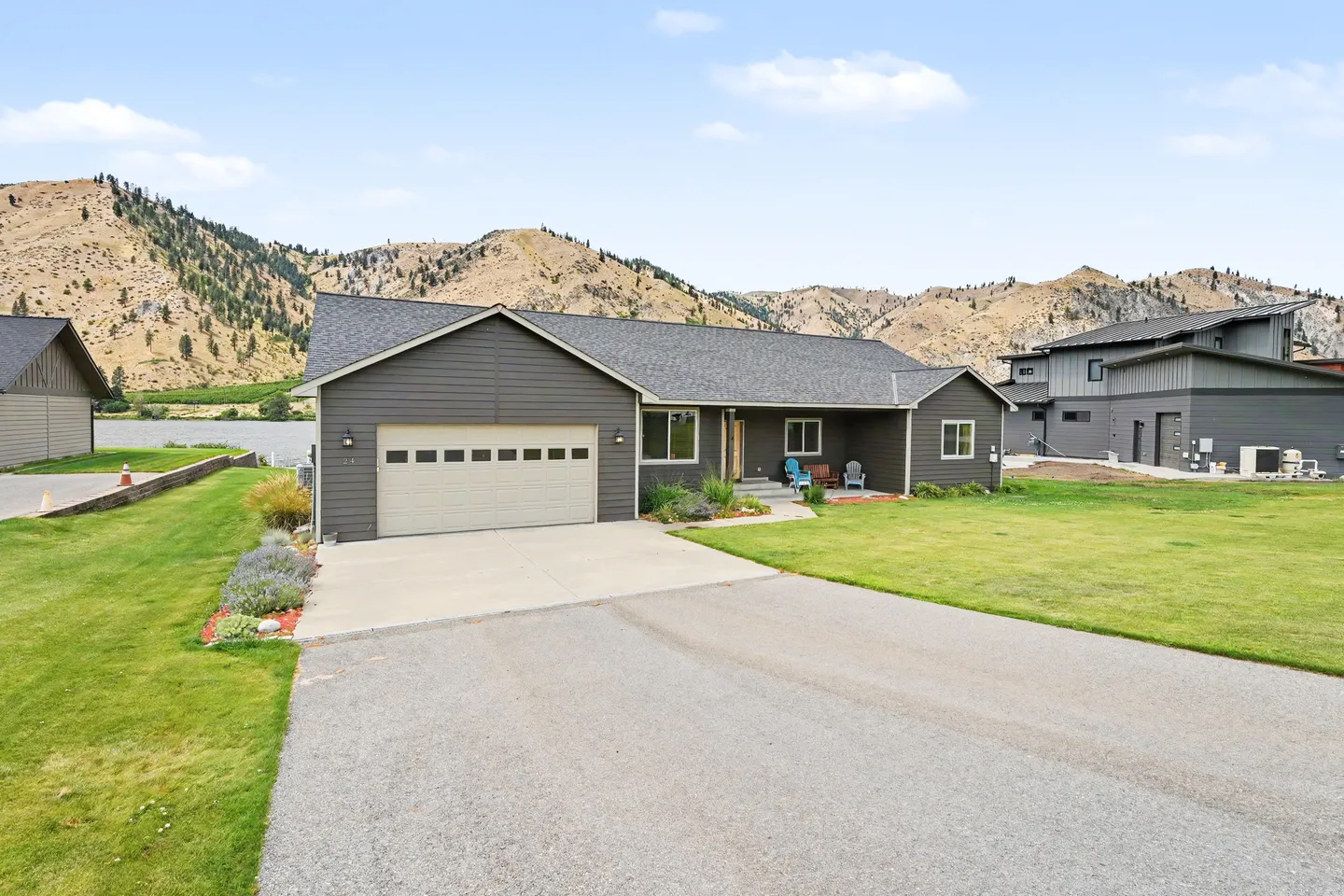 Exterior front view of a single-story dark siding house with an attached garage, green lawn, paved driveway, and mountain backdrop.