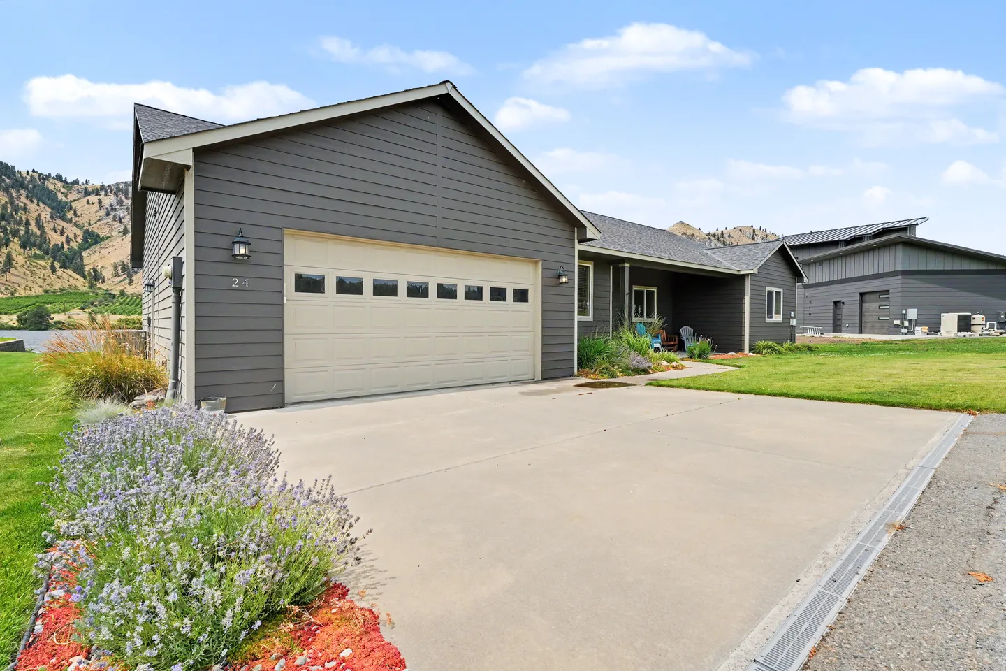 Exterior of a single-story gray home with an attached garage, wide concrete driveway, and front lawn with hills in the background.
