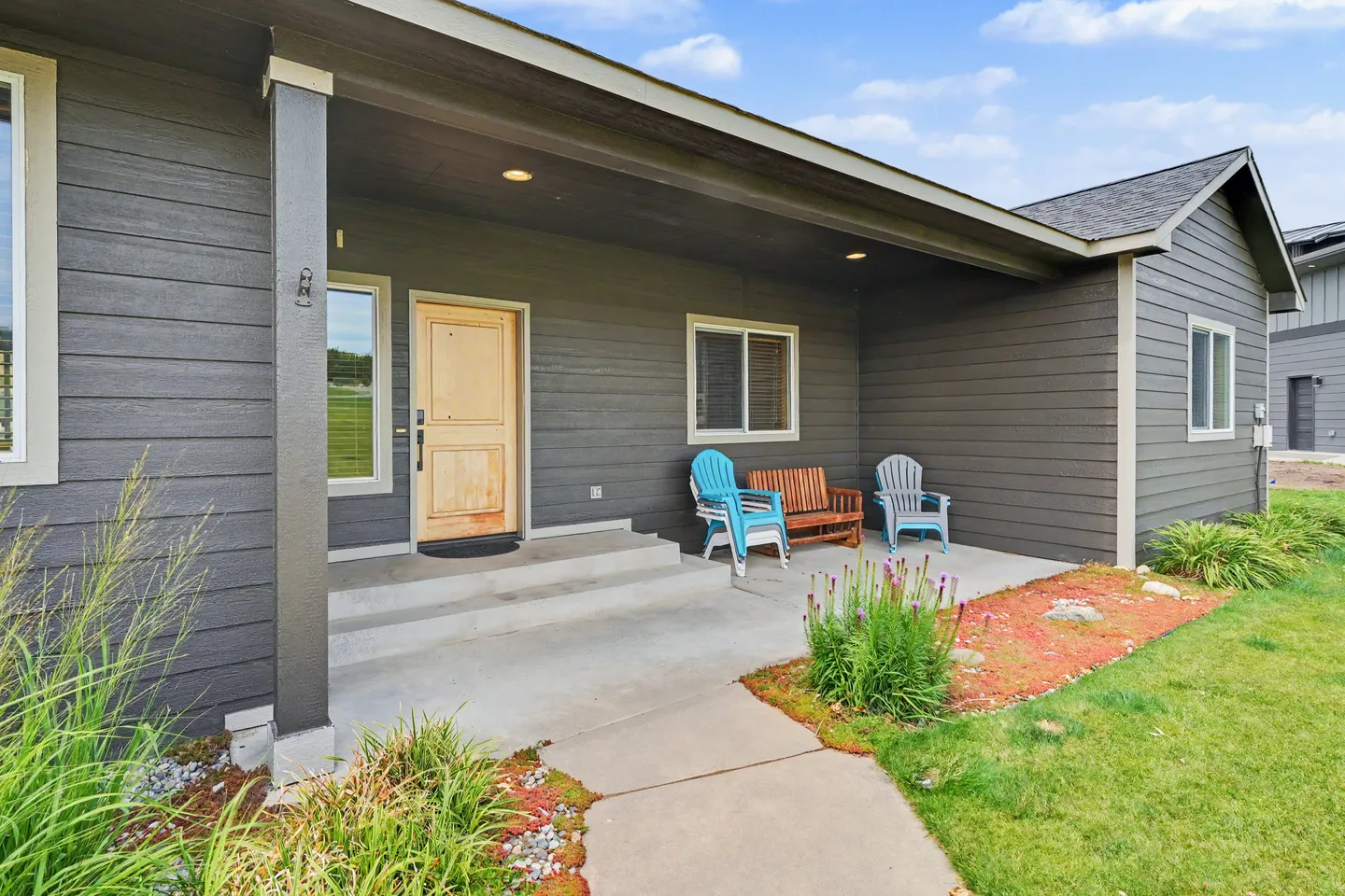 Front covered porch of a home with dark siding, a wooden front door, and seating including a bench and chairs.