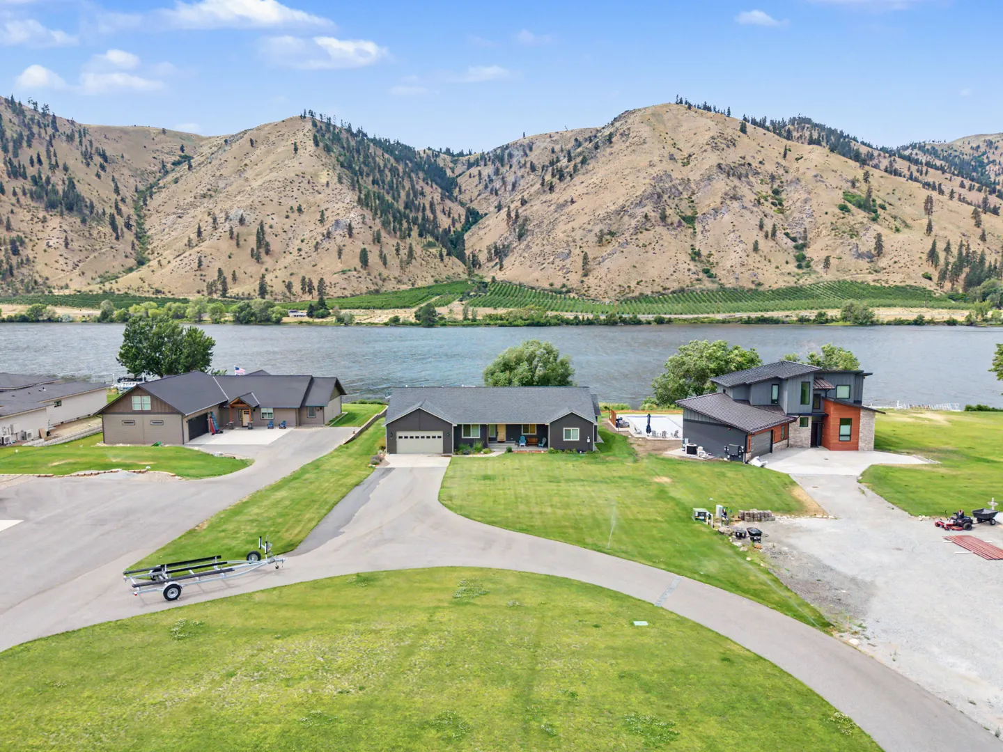 Aerial view of a waterfront home with a curved driveway situated along a river with mountains in the background.