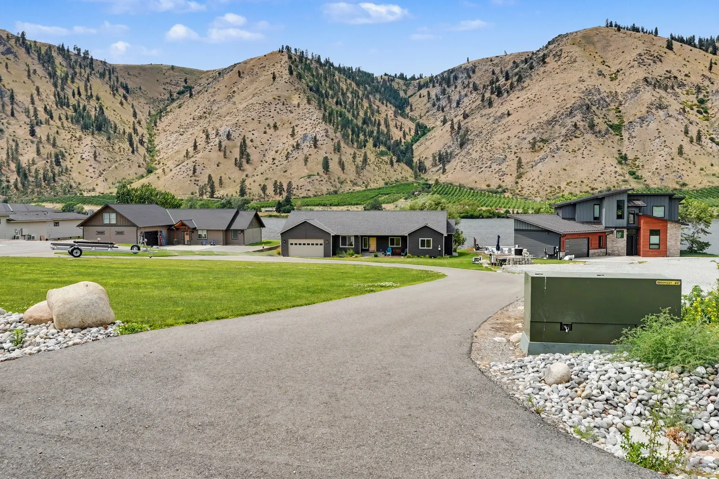A paved neighborhood road leading toward homes set against a lake and dry mountainous hills.