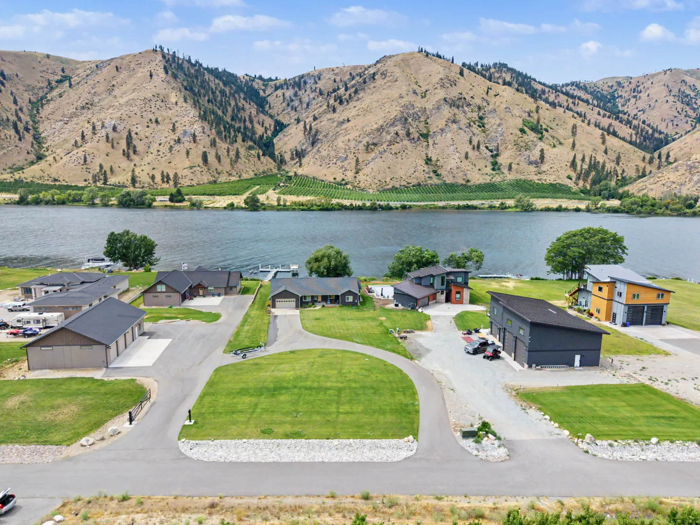 Aerial view of waterfront houses with green lawns along a river, backed by arid, mountainous terrain under a partly cloudy sky.