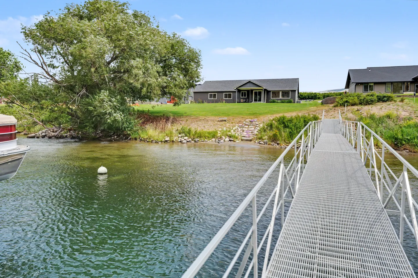 View from a metal dock walkway looking toward a grassy shoreline and a single-story house by the water.