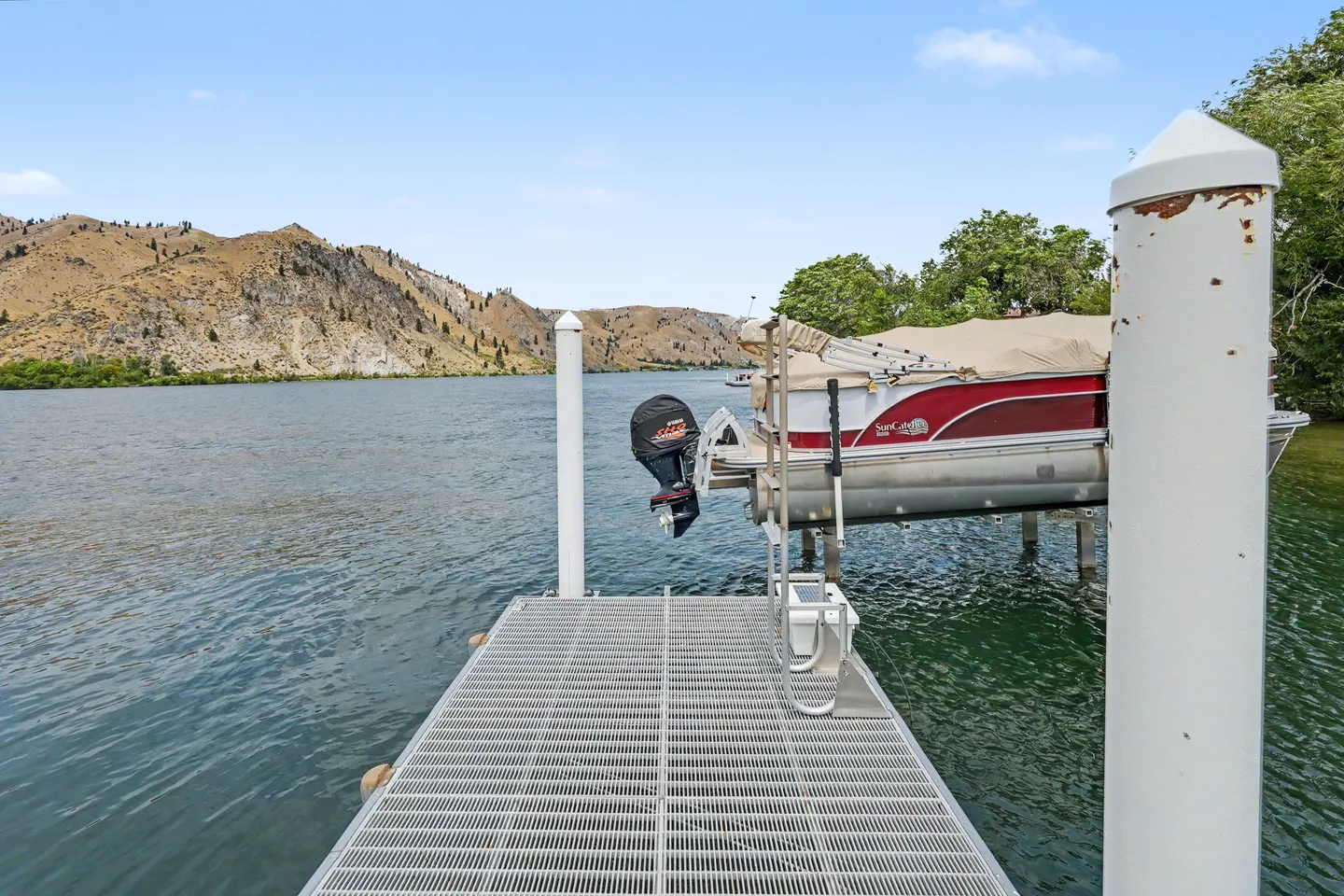 A metal dock extends into a lake toward hills, with a pontoon boat hoisted on a boat lift.