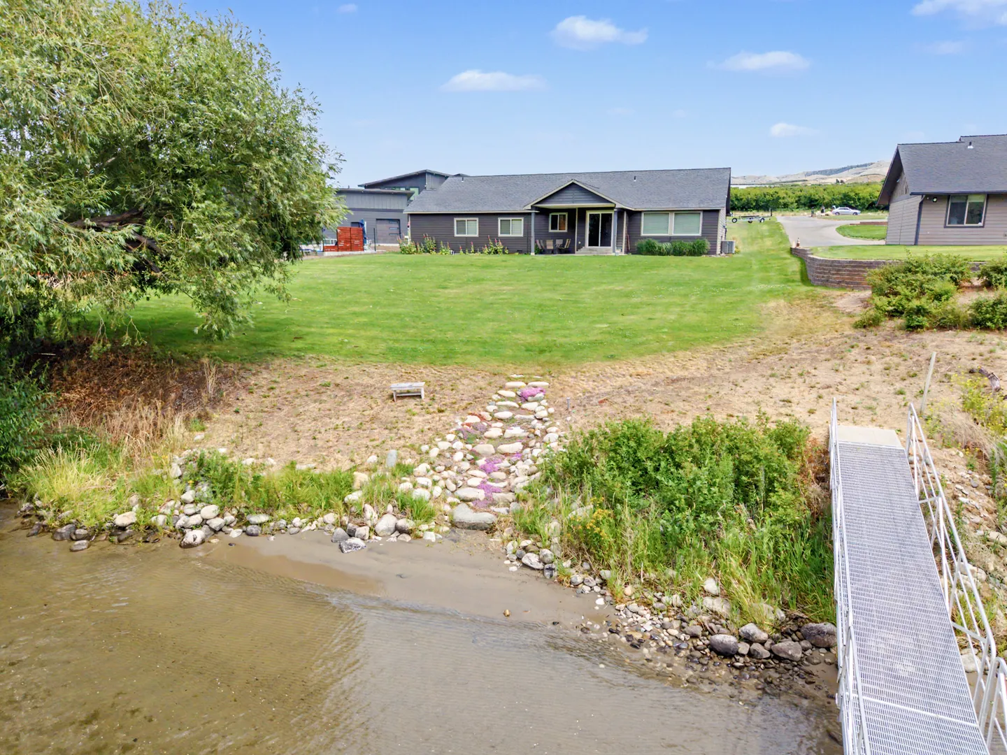 Waterfront view of a single-story dark-sided house with a large green lawn leading down to a rocky shore and metal ramp.