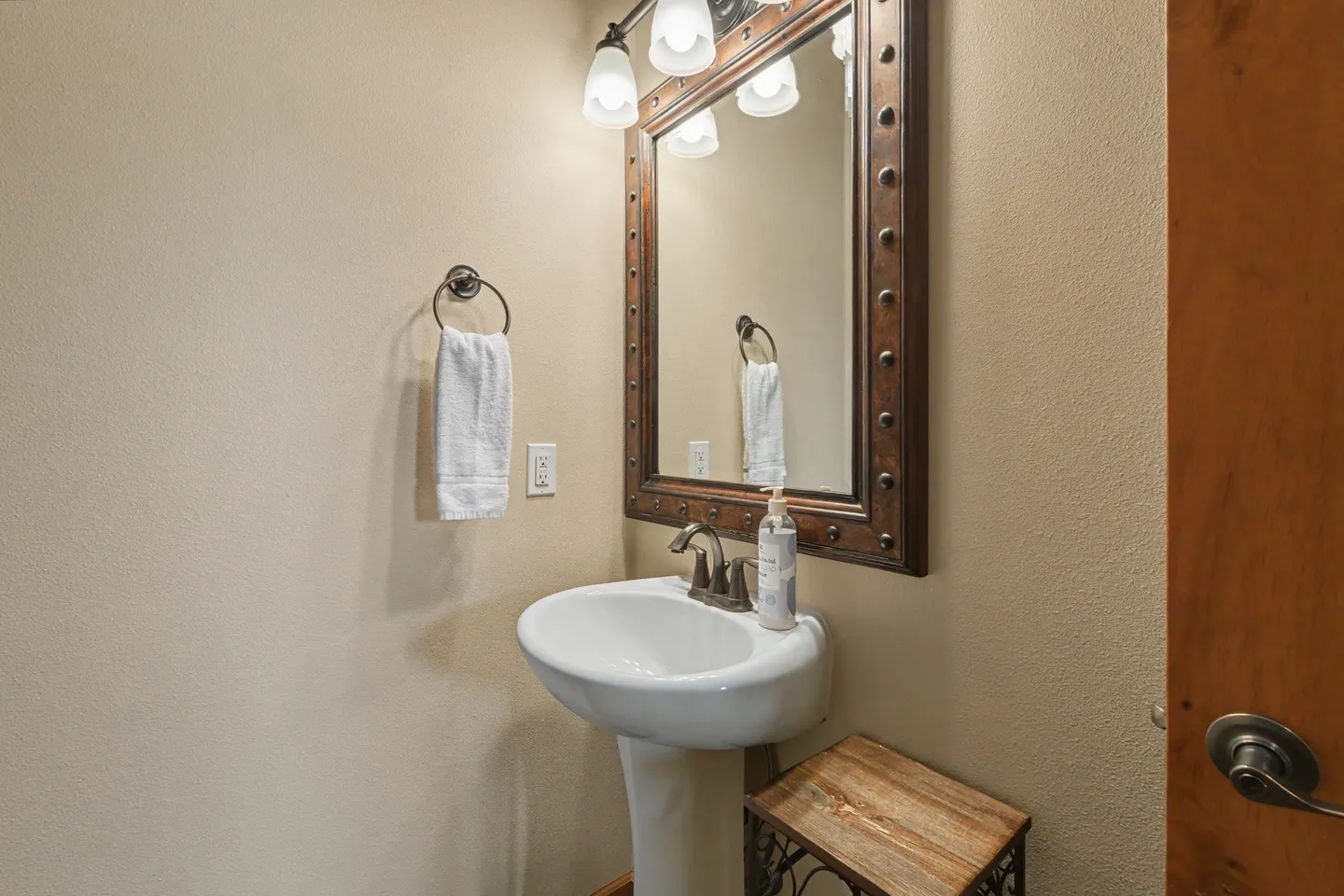 Powder room featuring a white pedestal sink, a framed mirror with metal stud accents, and overhead vanity lighting.