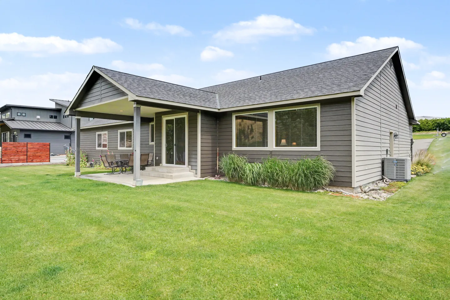 Rear exterior of a single-story house featuring horizontal siding, a covered concrete patio with outdoor seating, and a large green lawn under a partly cloudy sky.