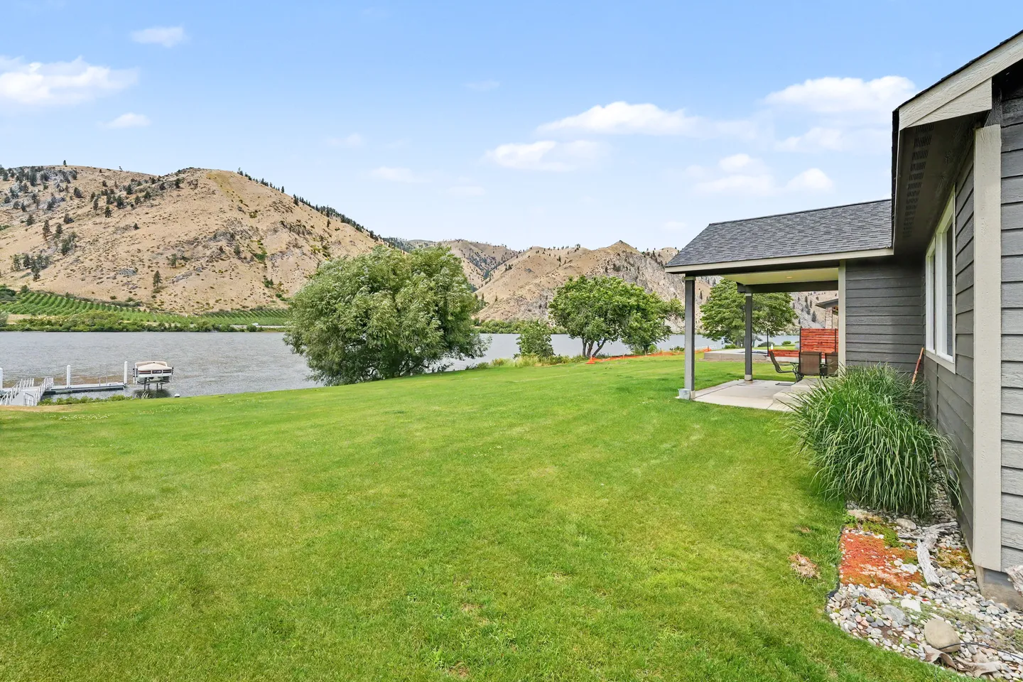 Lawn and covered patio behind a home overlooking a river and rugged hills with a boat dock visible on the water.