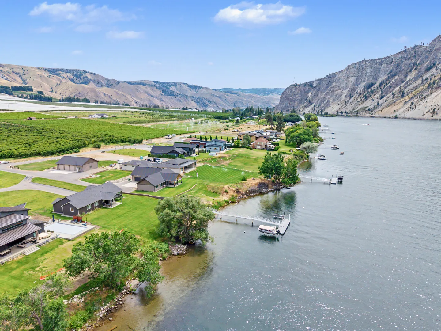 Aerial view of a waterfront neighborhood with houses, green lawns, private docks, and a wide river bordered by mountains and orchards.