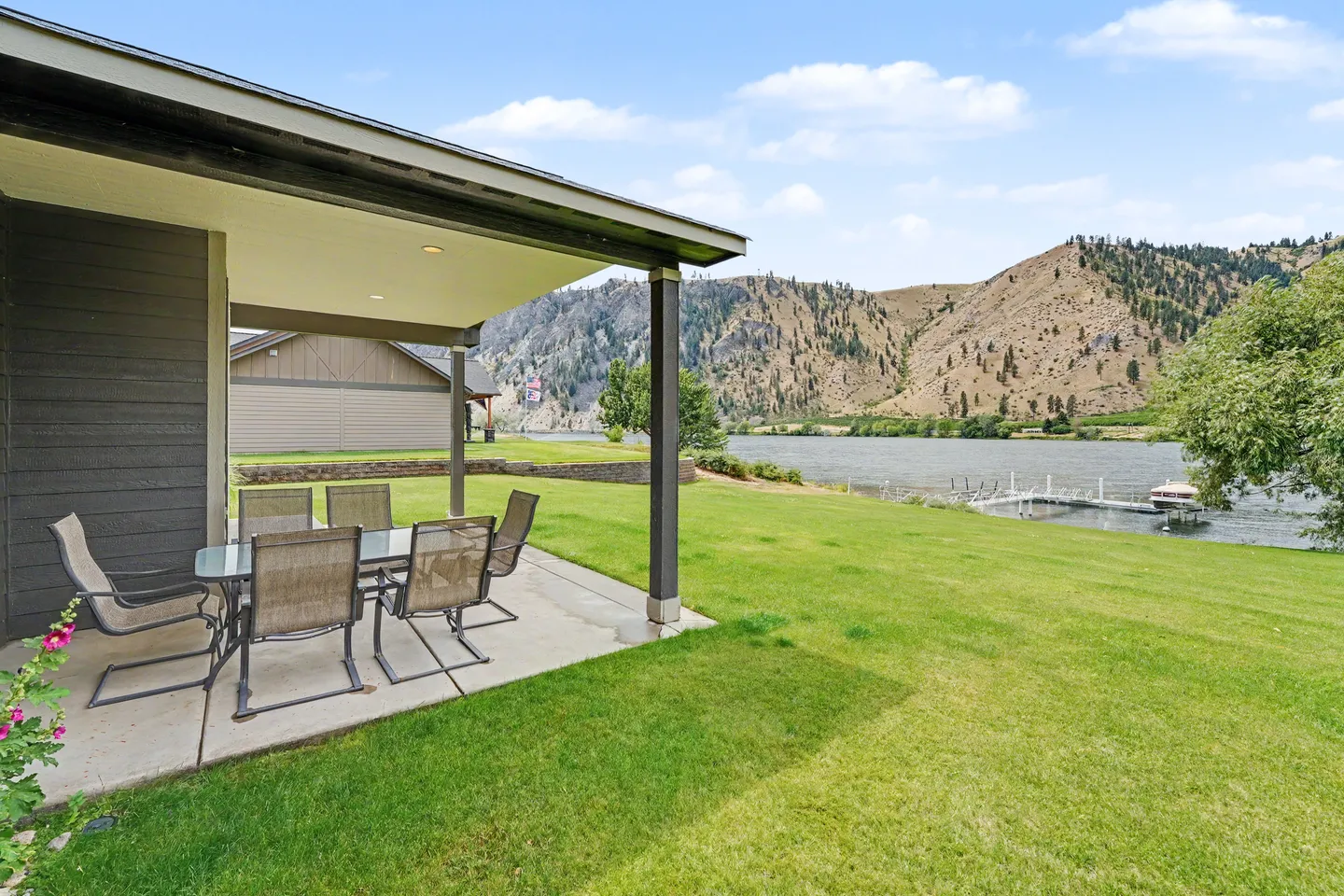 A covered concrete patio with outdoor dining furniture looking out over a grassy lawn toward a river and hills.