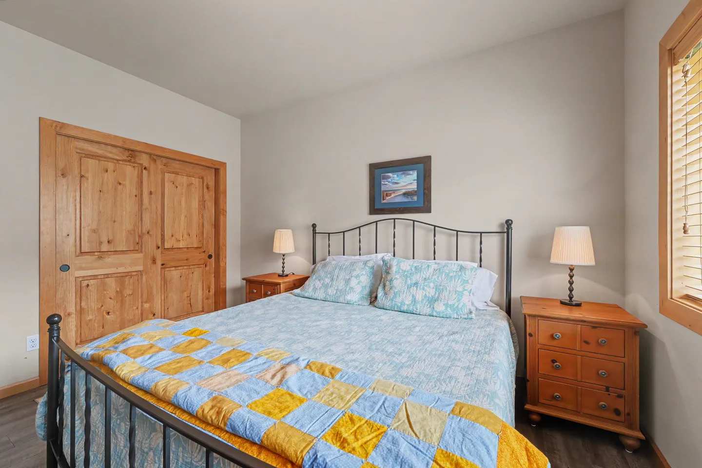 Bedroom featuring a black metal frame bed with blue and yellow patterned bedding, flanked by matching wood nightstands with lamps, near a wood closet door.