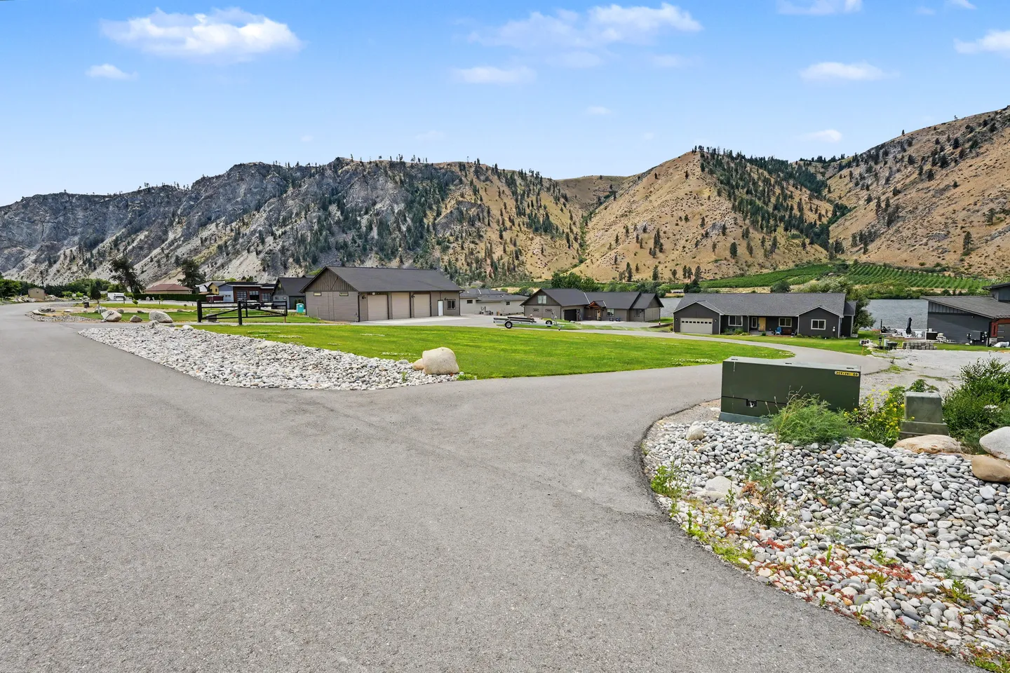 Paved neighborhood road leading past homes and manicured lawns toward mountains and water.