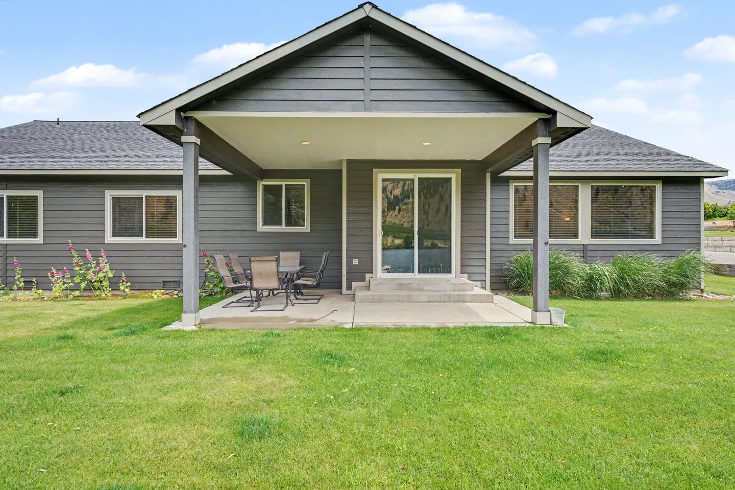 Exterior view of a dark gray house with a covered concrete patio, outdoor dining set, sliding glass door, and a green lawn.