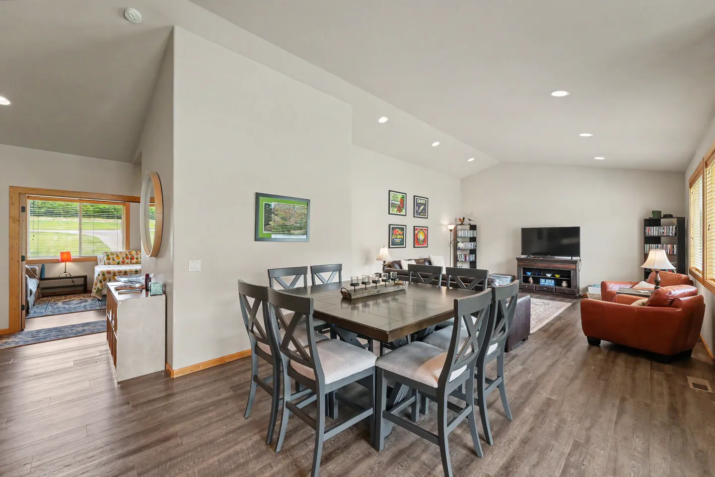 Open-concept dining and living area with a square dining table, grey chairs, wood-style flooring, and a view into an adjacent bedroom.