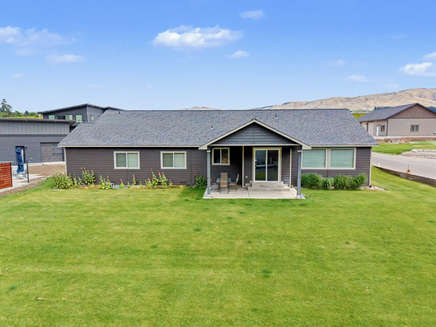 Exterior rear view of a single-story brown home with a covered patio and a large green lawn under a blue sky.