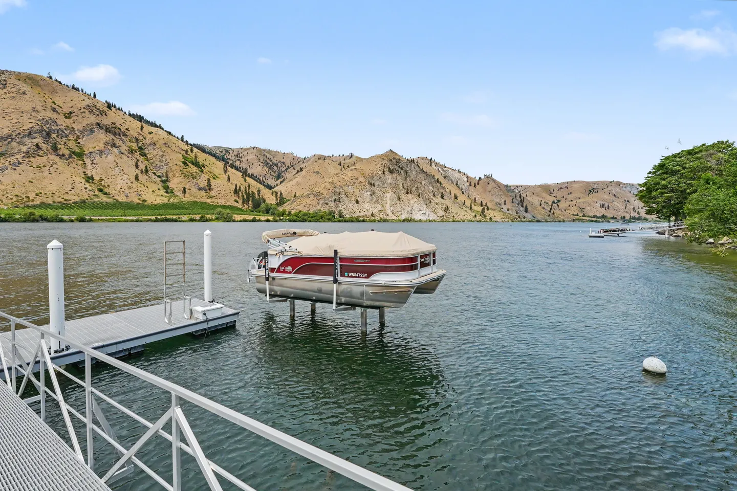A pontoon boat elevated on a boat lift next to a dock on a lake with arid hills in the background.