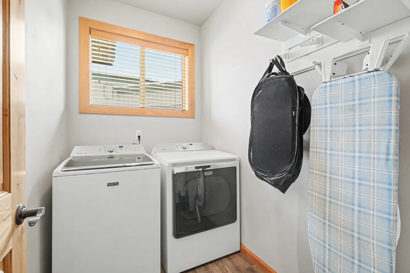 Laundry room with a white washing machine, white dryer, a window with horizontal blinds, and a mounted ironing board.